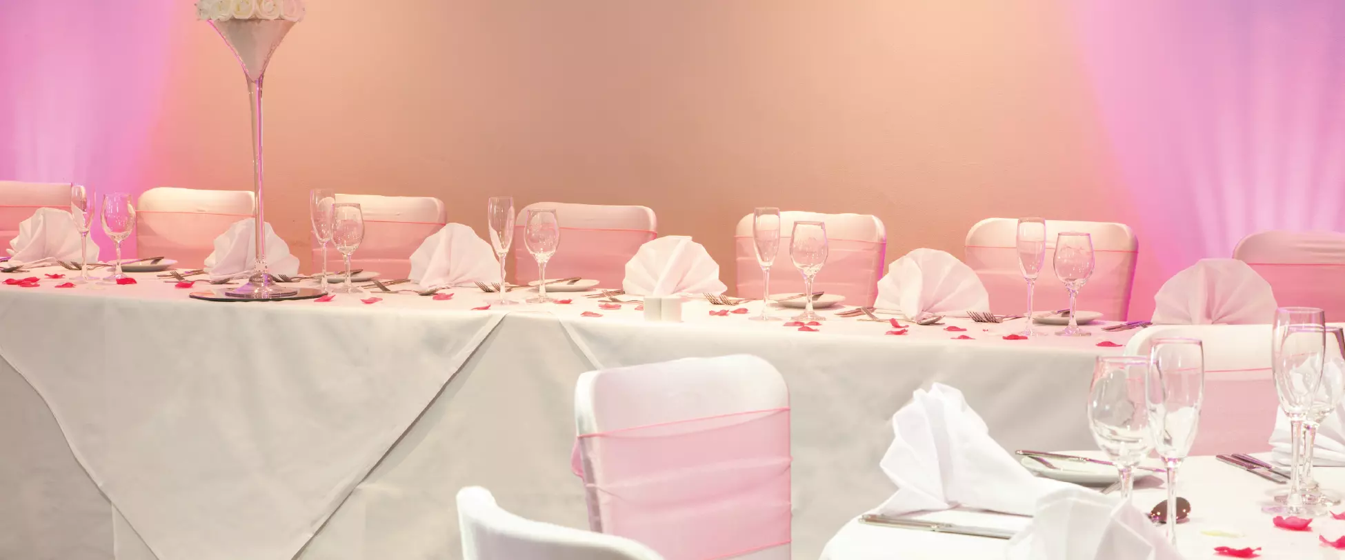 A table set up for the wedding party at a wedding breakfast. The table and chairs have white covers and the chairs have pink ribbons..